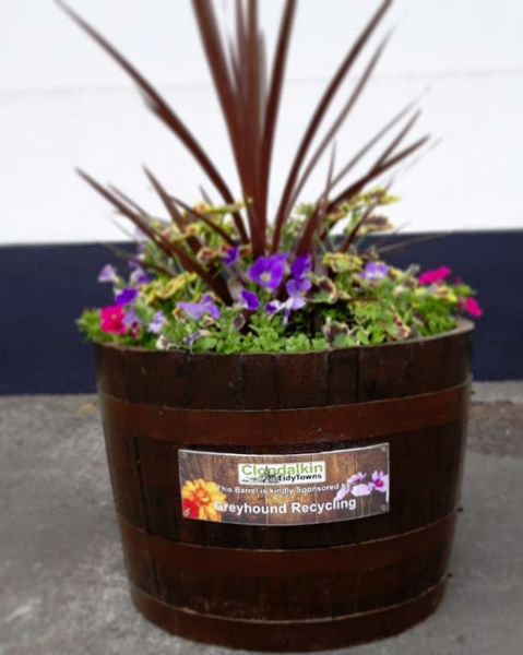 It shows the Greyhound logo and Clondalkin Tidy Towns logo on a wooden flower barrel.