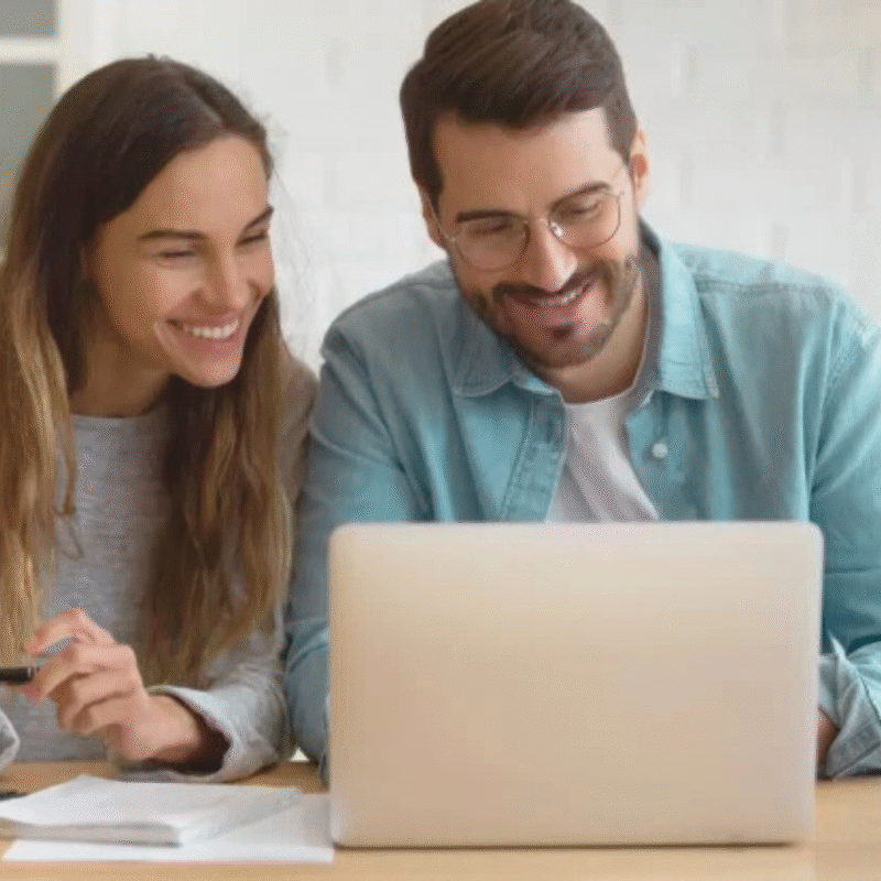 Husband and wife sitting in their kitchen looking at their laptop. They have notes in front of them and the wife has a pen in hand. They are malking a decision about something.