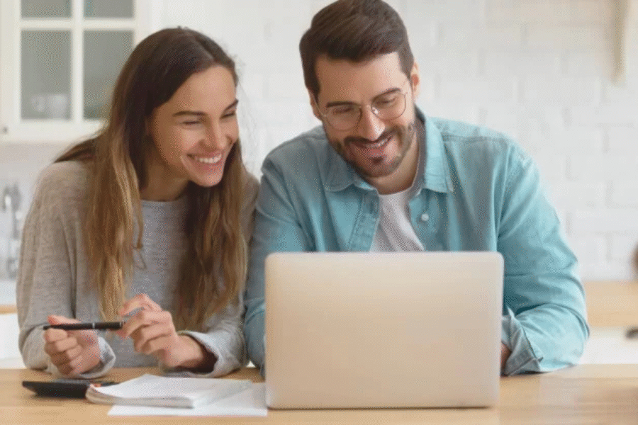 Husband and wife sitting in their kitchen looking at their laptop. They have notes in front of them and the wife has a pen in hand. They are malking a decision about something.